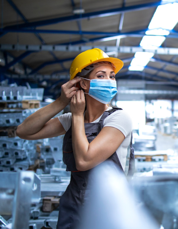 Shot of female factory worker in uniform and hardhat putting on face mask in industrial production plant.